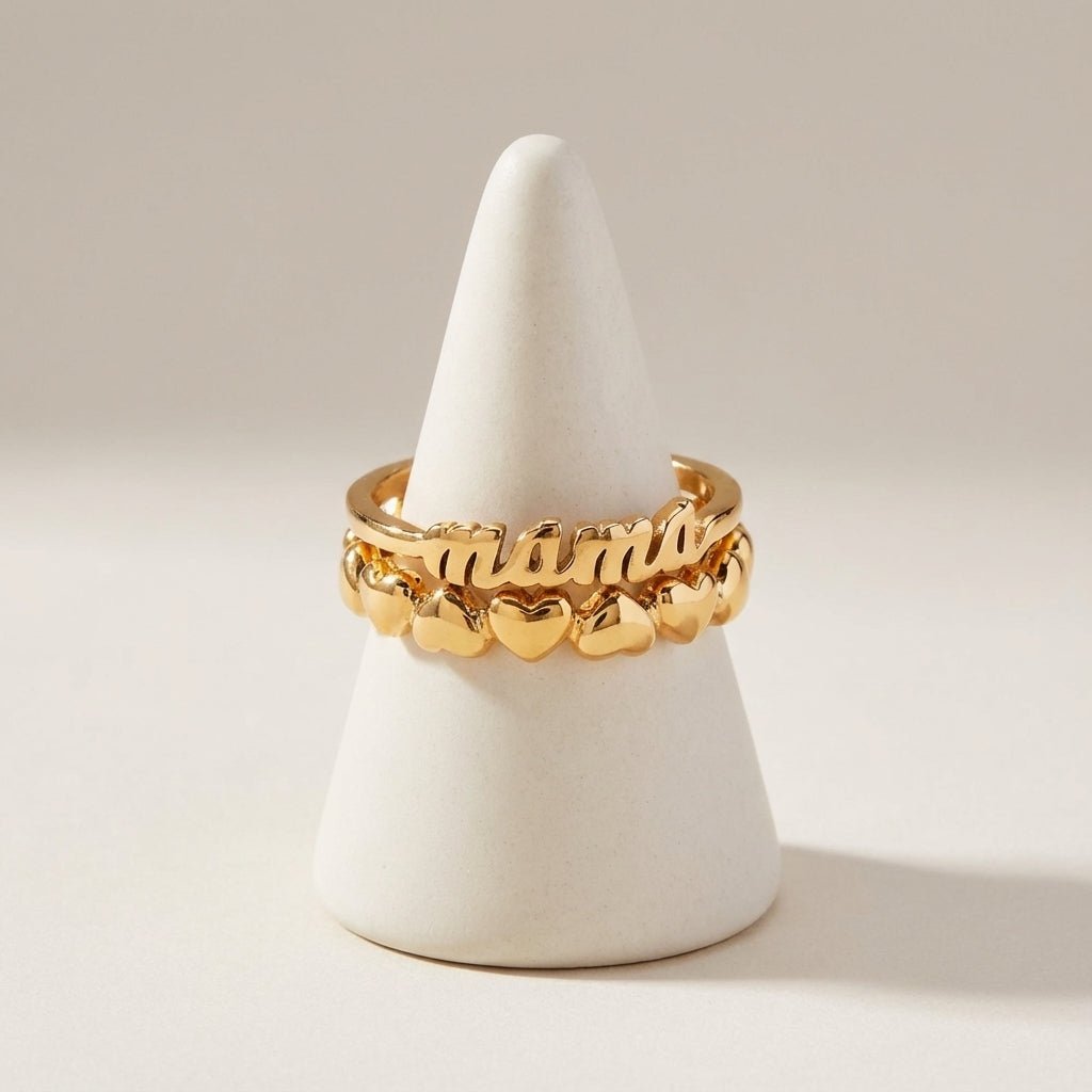 Gold ring with 'mama' and heart designs on a white stand against a beige background