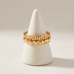Gold ring with 'mama' and heart designs on a white stand against a beige background