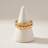 Gold ring with 'mama' and heart designs on a white stand against a beige background
