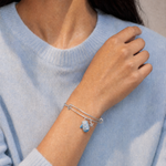 A close-up of a person wearing a delicate silver wire charm bracelet with a small blue sea otter charm, paired with a light blue knit sweater.