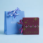 A light blue Alex + Ani gift bag with a blue and white patterned ribbon next to a dark burgundy gift box with green star patterns and a pink patterned band, both printed with the brand name 'ALEX + ANI', on a light blue background.