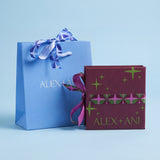 A light blue Alex + Ani gift bag with a blue and white patterned ribbon next to a dark burgundy gift box with green star patterns and a pink patterned band, both printed with the brand name 'ALEX + ANI', on a light blue background.