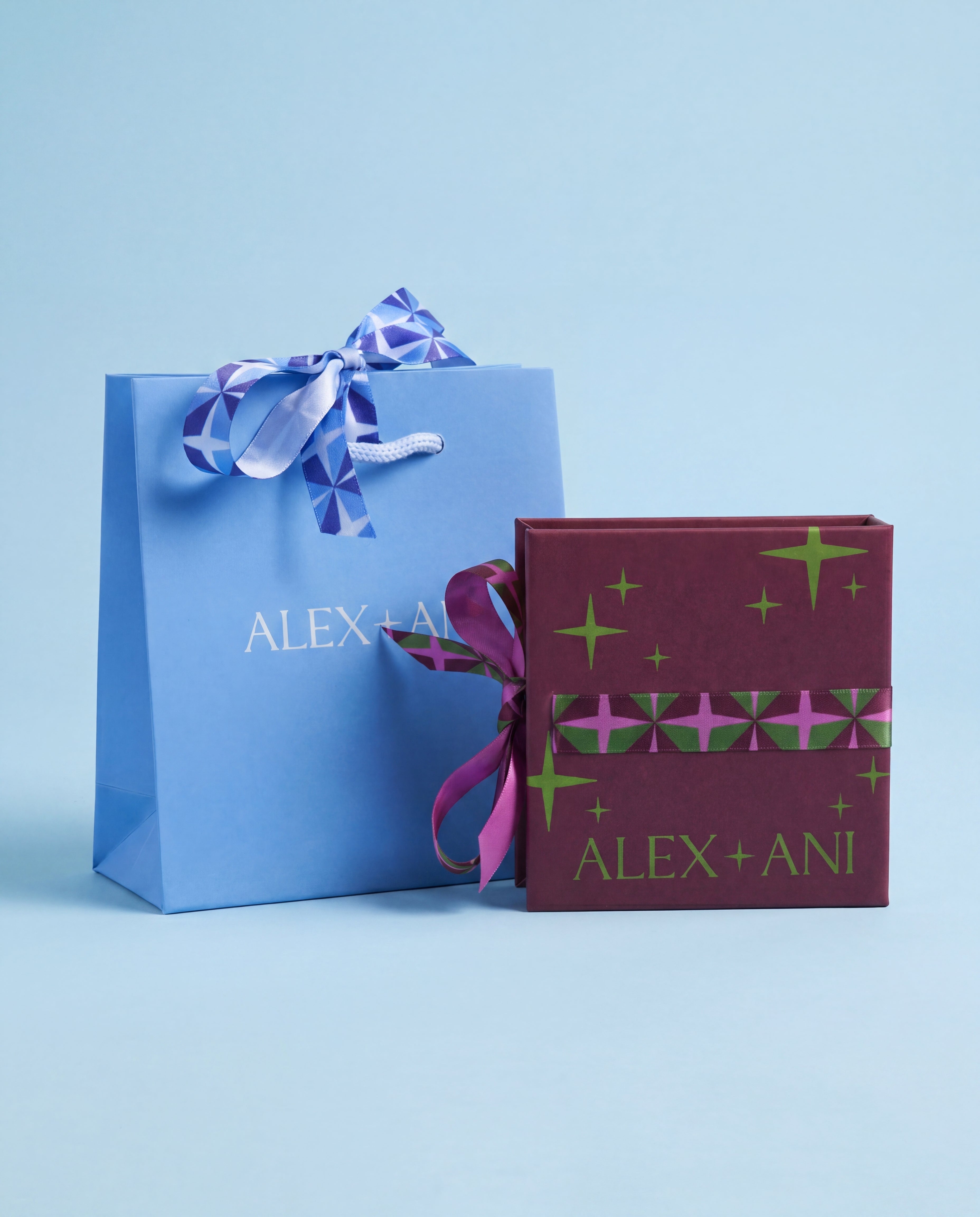A light blue Alex + Ani gift bag with a blue and white patterned ribbon next to a dark burgundy gift box with green star patterns and a pink patterned band, both printed with the brand name 'ALEX + ANI', on a light blue background.