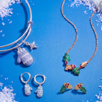 A jewelry flat-lay featuring a silver bangle bracelet, a gold necklace, and matching silver and gold hoop earrings. The jewelry is decorated with a light blue snowflake-patterned mitten charm, a small star charm, and a holly-and-mistletoe motif with red berries, all arranged on a bright blue background with faux snow accents.