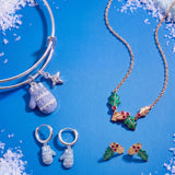 A jewelry flat-lay featuring a silver bangle bracelet, a gold necklace, and matching silver and gold hoop earrings. The jewelry is decorated with a light blue snowflake-patterned mitten charm, a small star charm, and a holly-and-mistletoe motif with red berries, all arranged on a bright blue background with faux snow accents.