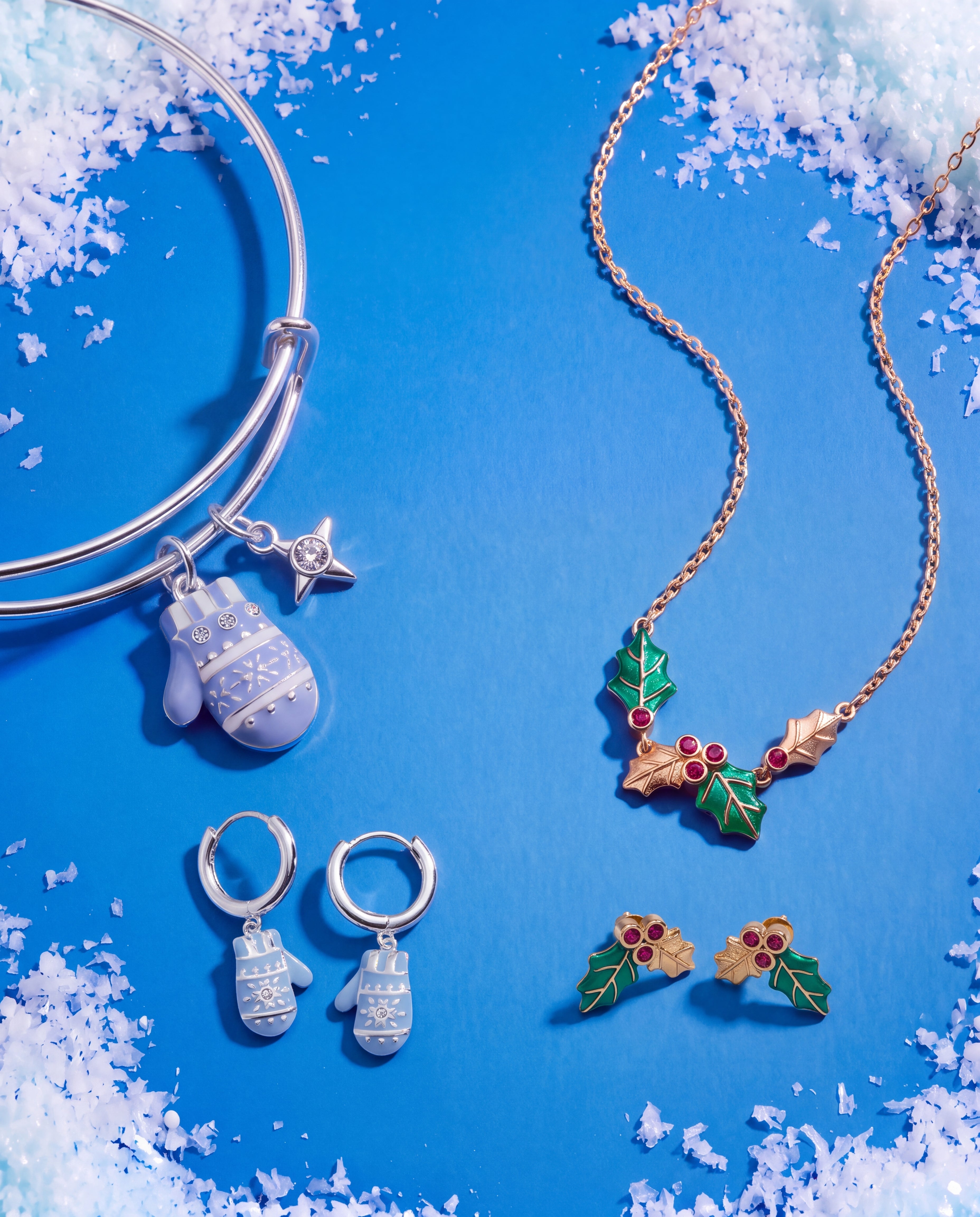 A jewelry flat-lay featuring a silver bangle bracelet, a gold necklace, and matching silver and gold hoop earrings. The jewelry is decorated with a light blue snowflake-patterned mitten charm, a small star charm, and a holly-and-mistletoe motif with red berries, all arranged on a bright blue background with faux snow accents.