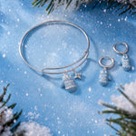 A silver bangle bracelet and matching drop earrings with snowflake and star charms, styled on a blue snowy background with pine branches.