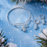 A silver bangle bracelet and matching drop earrings with snowflake and star charms, styled on a blue snowy background with pine branches.