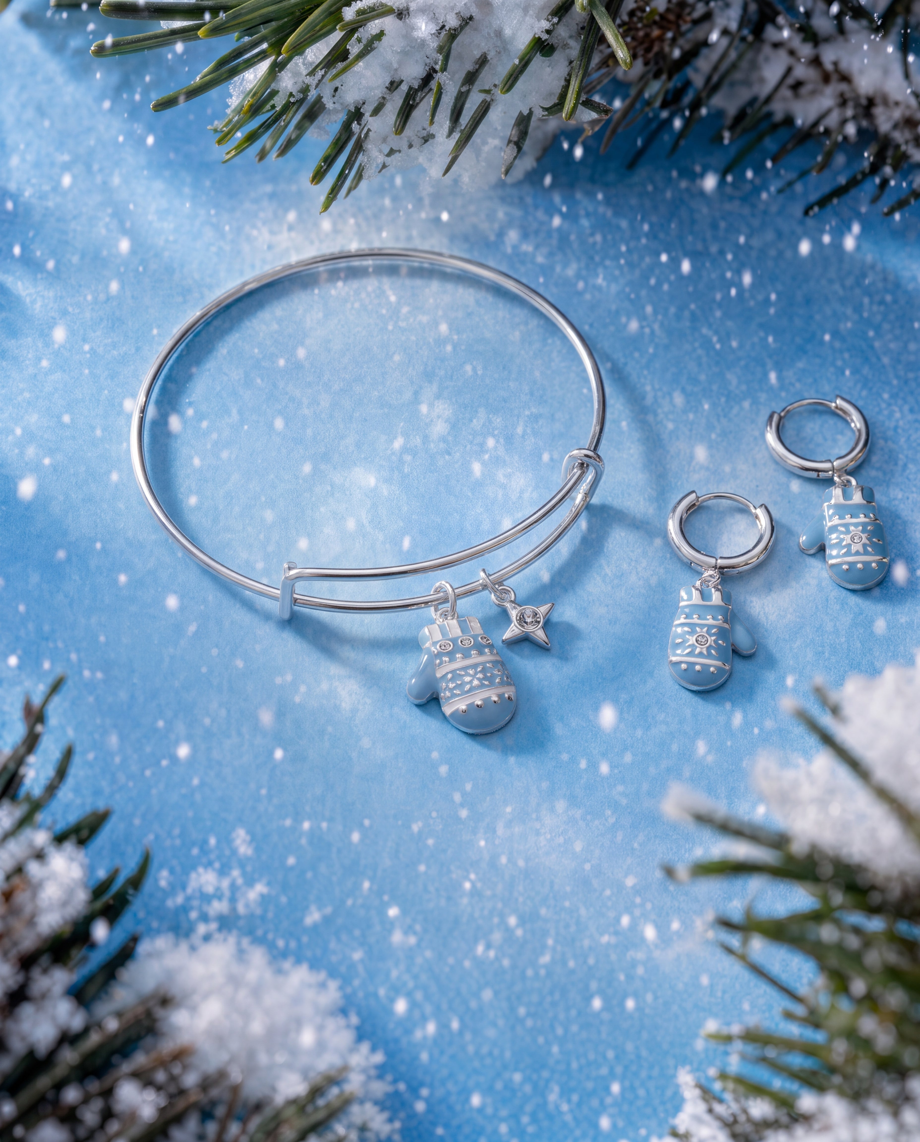 A silver bangle bracelet and matching drop earrings with snowflake and star charms, styled on a blue snowy background with pine branches.