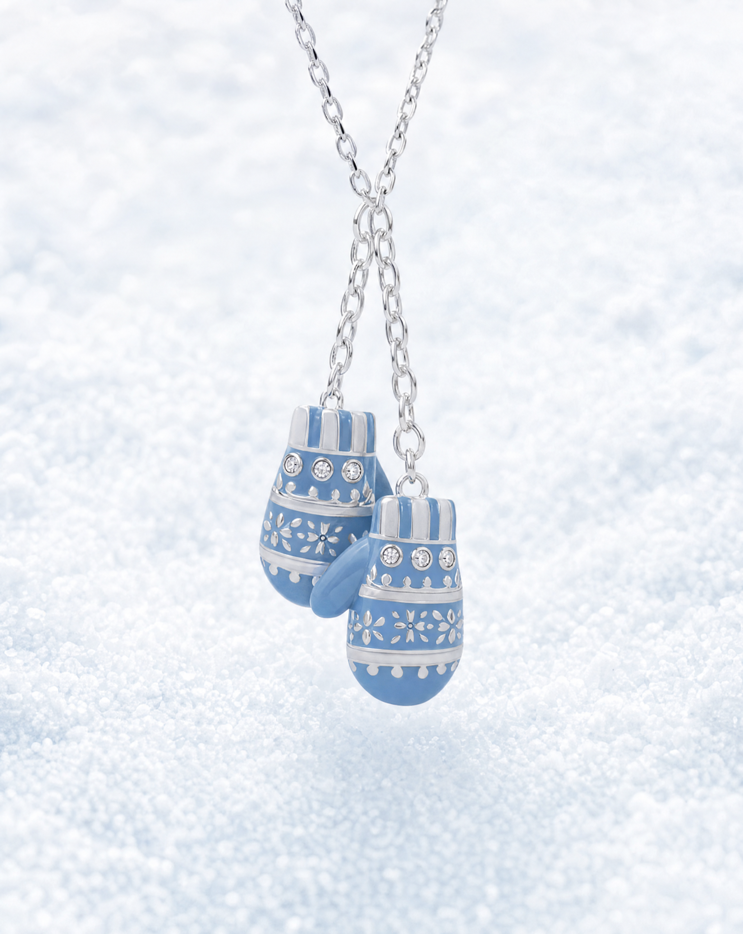 A silver chain necklace with a pair of blue and white patterned mitten-shaped charms, photographed against a soft, snowy white background.