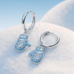 A pair of silver hoop earrings with dangling light blue mitten charms decorated with white snowflake patterns, resting on a bed of white snow with a soft blue sky background.