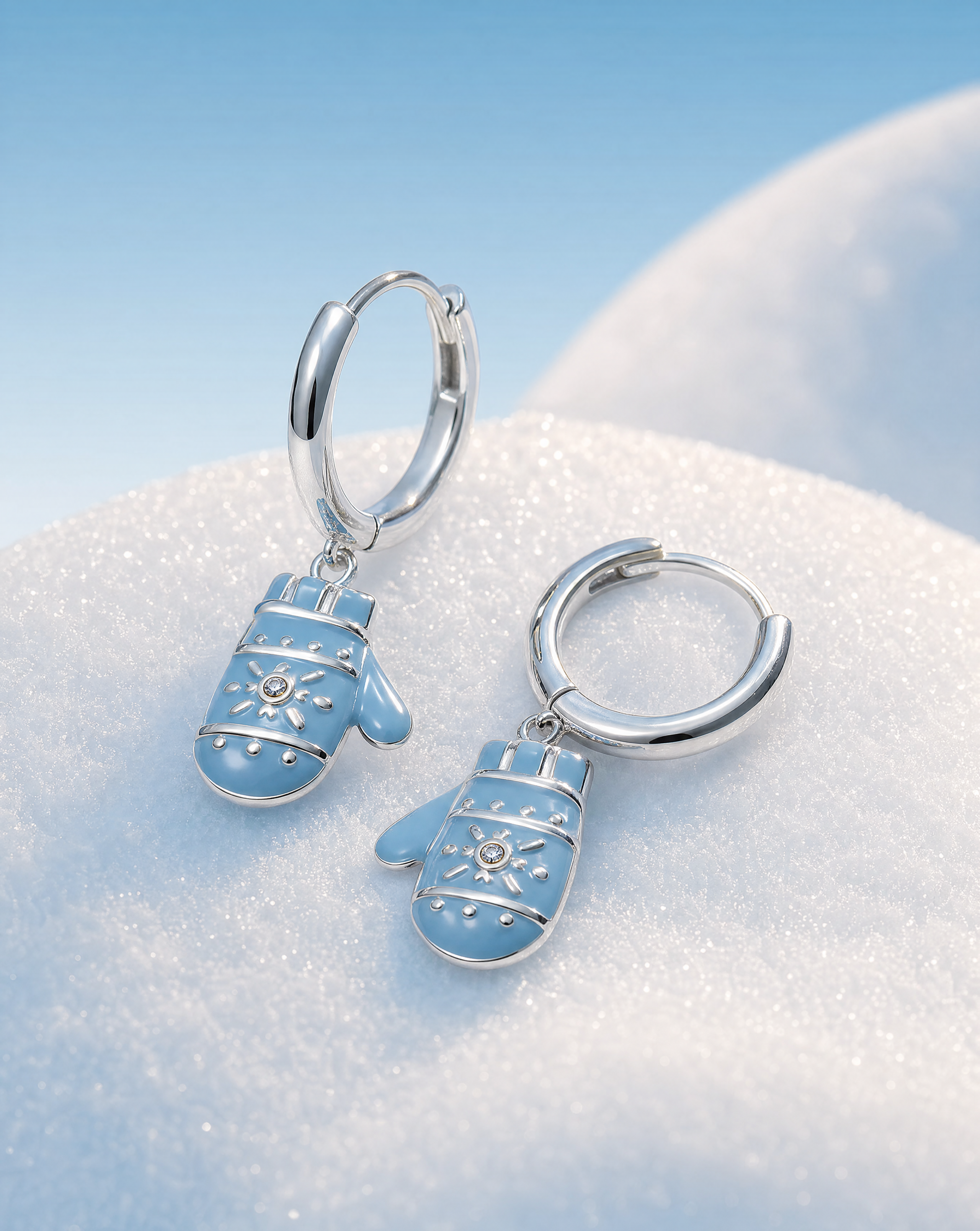 A pair of silver hoop earrings with dangling light blue mitten charms decorated with white snowflake patterns, resting on a bed of white snow with a soft blue sky background.