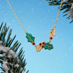A gold holly necklace with green enamel leaves and red berry accents, shown hanging against a bright blue sky background with snowy pine branches.