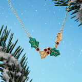 A gold holly necklace with green enamel leaves and red berry accents, shown hanging against a bright blue sky background with snowy pine branches.