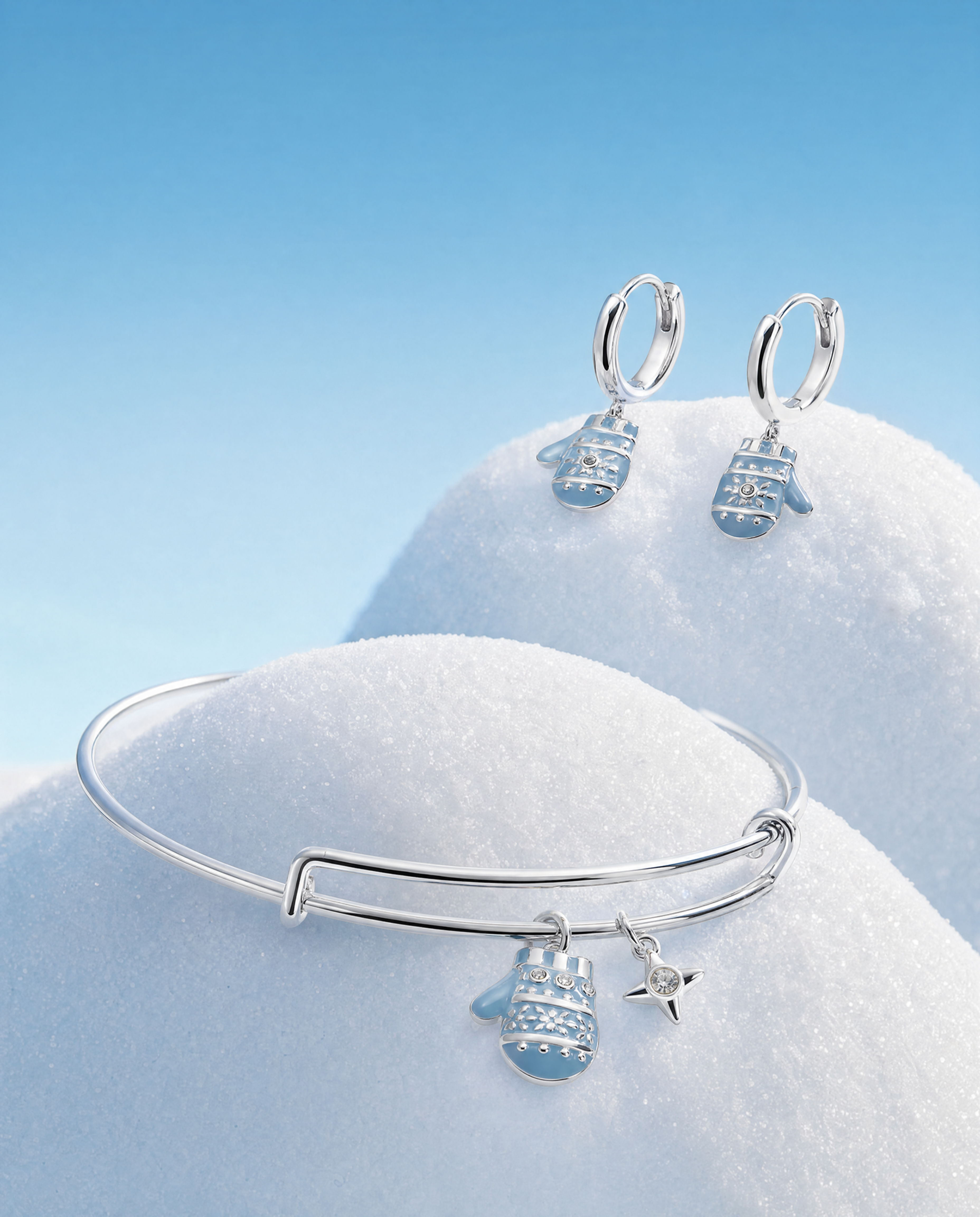 A silver bangle bracelet and matching drop earrings with snowflake and star charms, styled on a blue snowy background on top of two mounds of snow