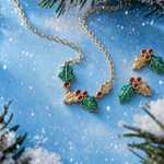 A necklace and a pair of matching gold-tone holly leaf stud earrings with vibrant green enamel leaves and three small round pink gemstones at the top, set against a snowy background with branches