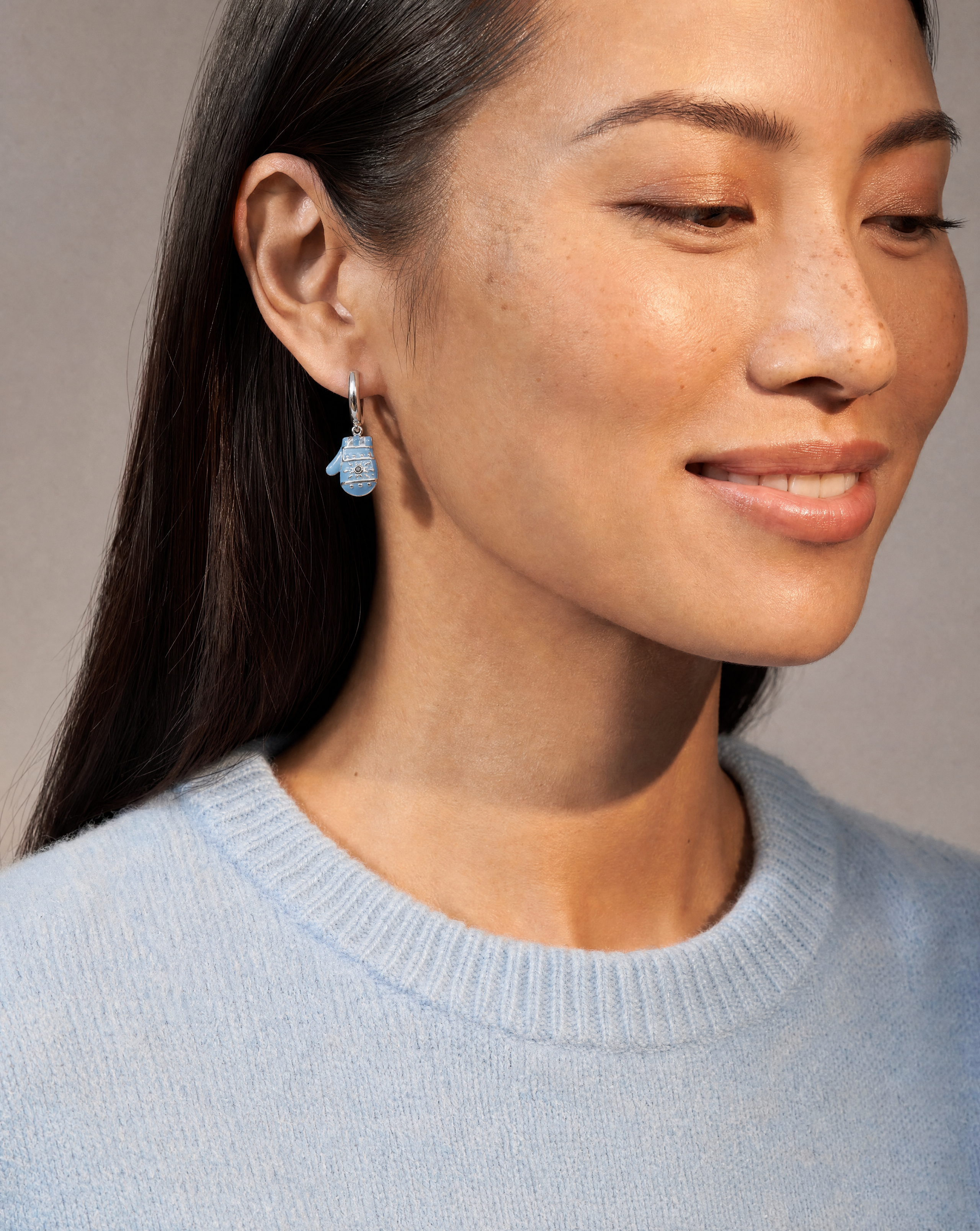 A close-up of a woman wearing a small light blue hamsa hand dangle earring with a silver hook, paired with a light blue knit sweater against a neutral gray background.