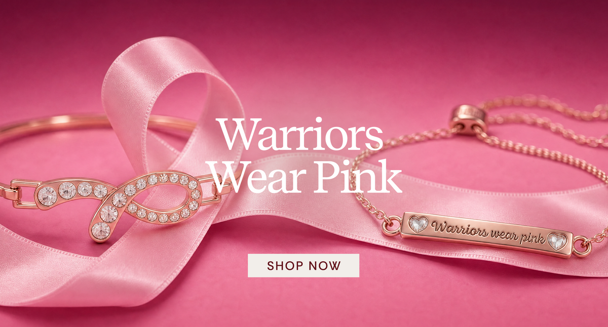 Rose gold jewelry set featuring a ribbon-shaped charm with clear crystals and a bar engraved with 'Warriors wear pink', displayed on a pink satin ribbon over a pink background with the text 'Warriors Wear Pink' and a 'SHOP NOW' button.