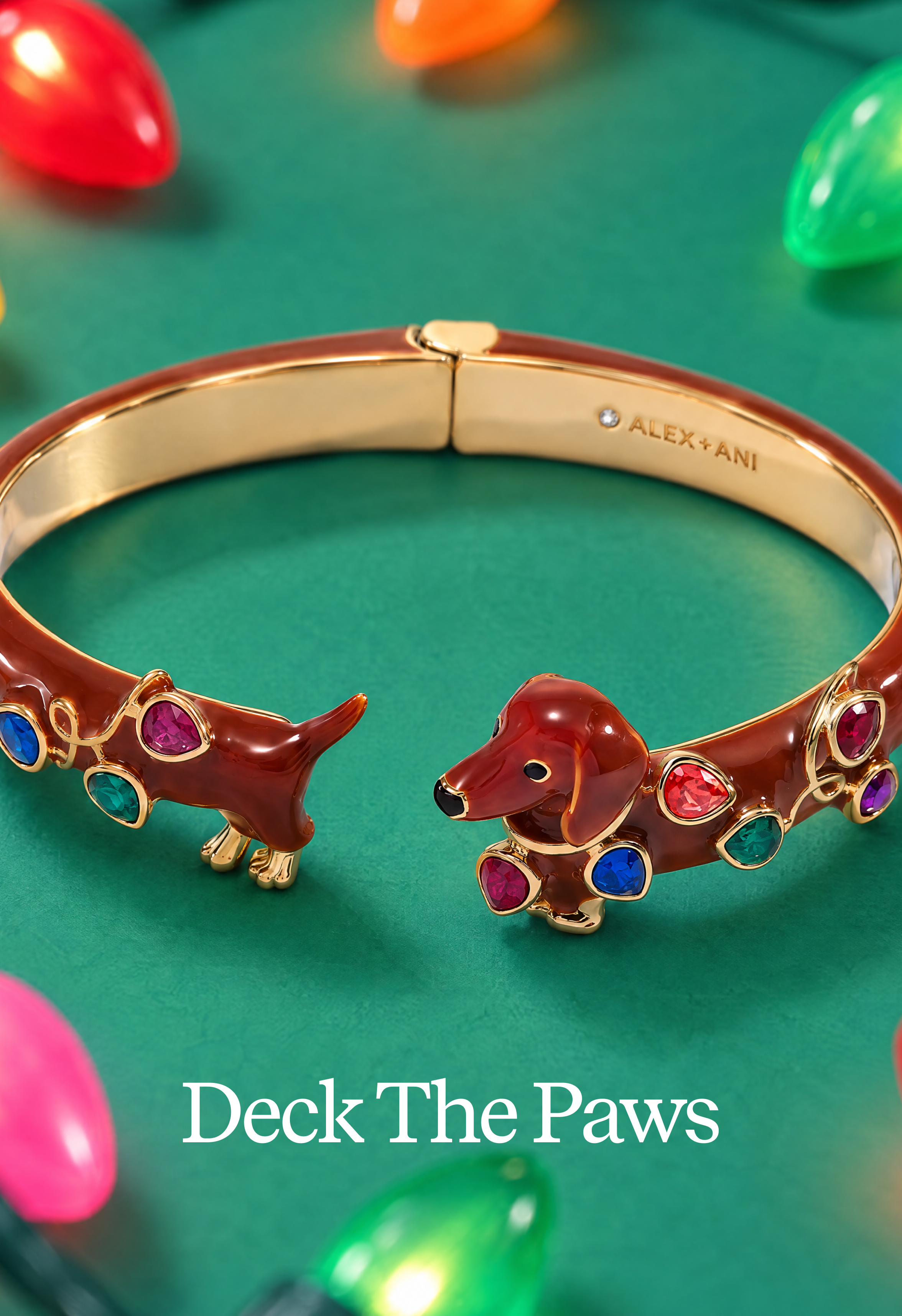 Gold-tone cuff bracelet with a dachshund-shaped end and colorful gem accents, photographed on a green surface with Christmas lights in the background. The inside of the bracelet is engraved with ALEX+ANI, and the text 'Deck The Paws' appears at the bottom.