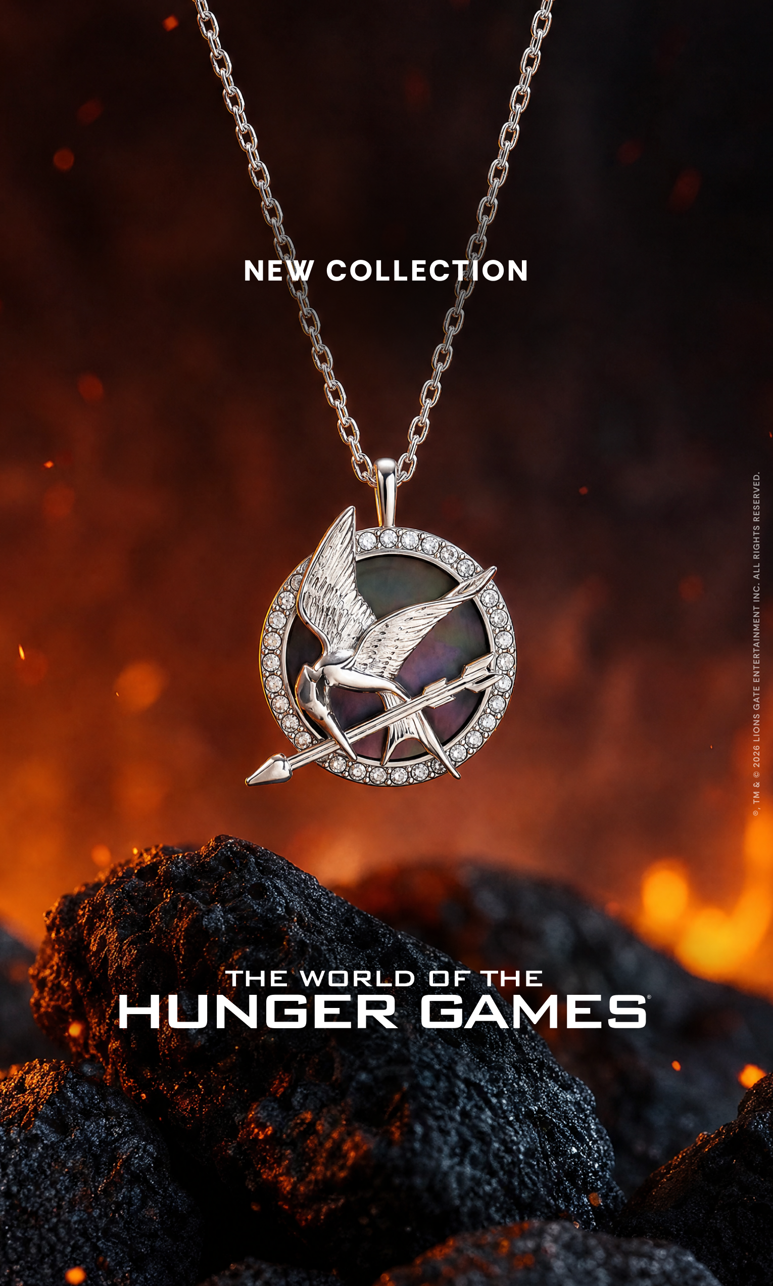 A silver-tone Hunger Games pendant necklace with a circular pin featuring a bird and arrow, displayed against a fiery black background with glowing embers and coals. Text on the image reads 'NEW COLLECTION' and 'THE WORLD OF THE HUNGER GAMES'.