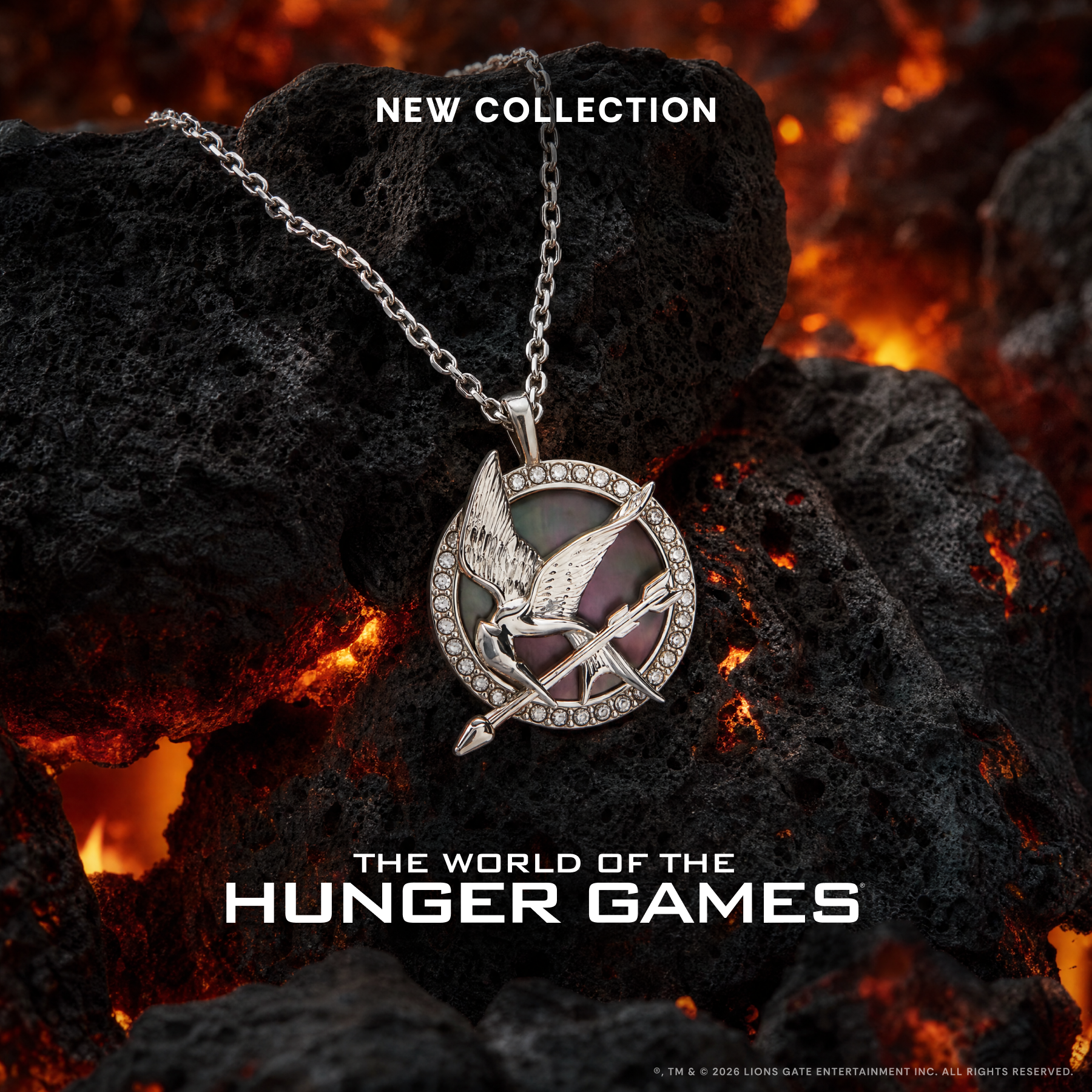 A silver-tone Hunger Games pin necklace with a mother-of-pearl patterned center and crystal-studded border, resting on dark volcanic rocks with glowing embers. Text overlays read 'NEW COLLECTION' and 'THE WORLD OF THE HUNGER GAMES'.