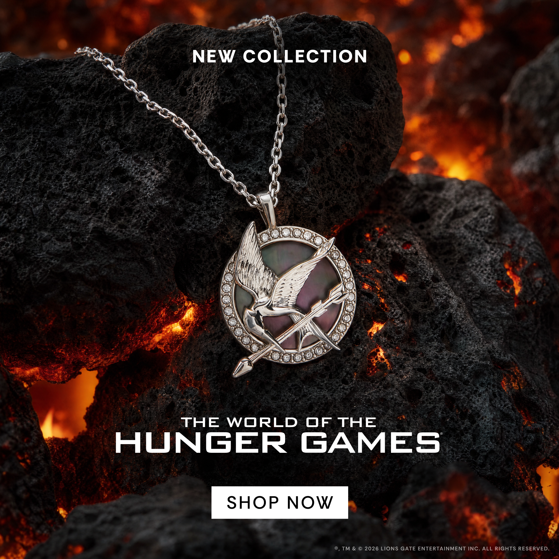 A silver-toned Hunger Games pendant necklace with a bird emblem and small clear stones, displayed on dark volcanic rocks with glowing embers. Text reads 'NEW COLLECTION', 'THE WORLD OF THE HUNGER GAMES', and 'SHOP NOW'.