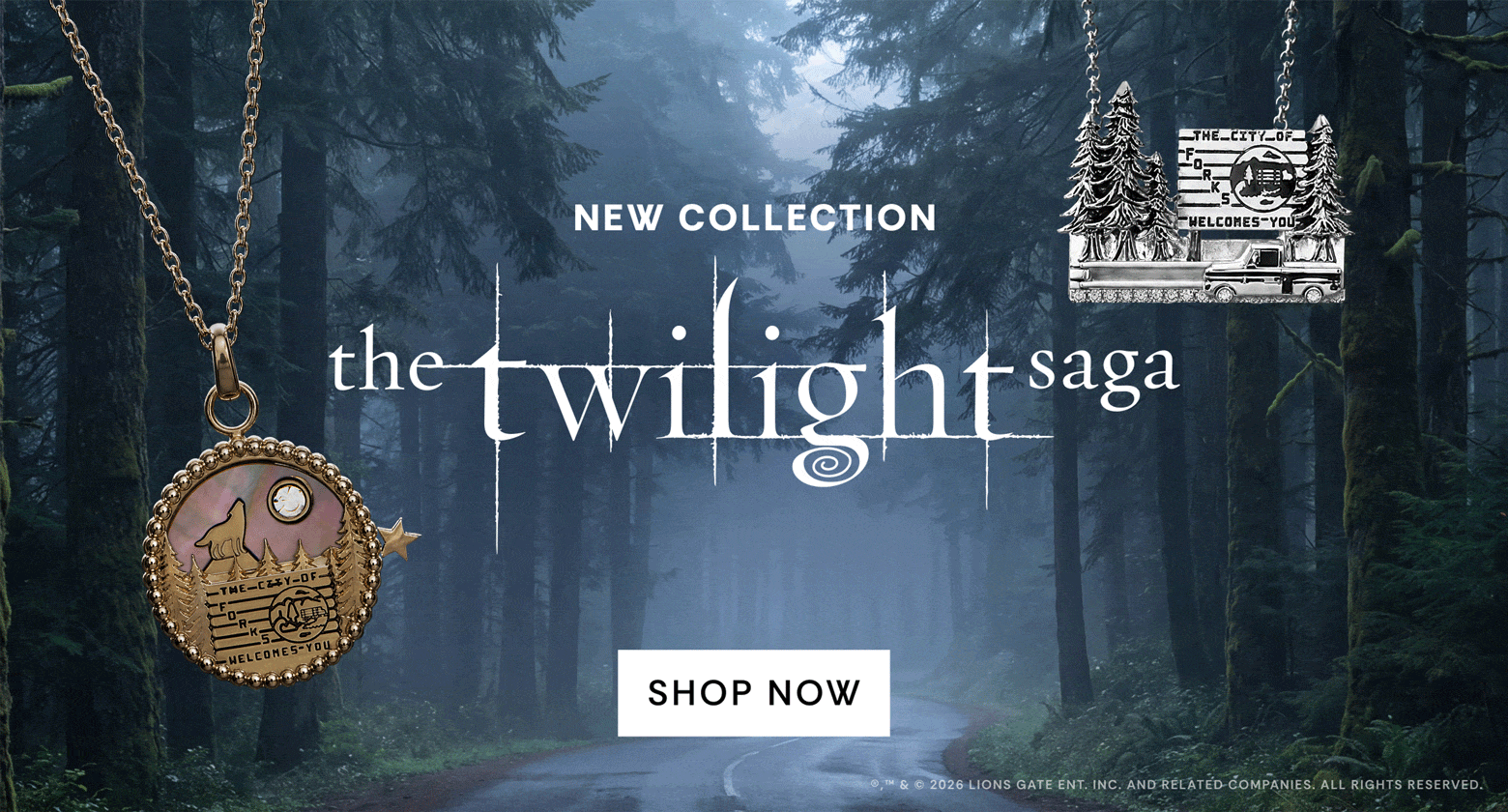 Promotional banner for 'The Twilight Saga' jewelry collection, featuring a silver pendant with a moon and stars design on the left and a bronze-toned pendant with a cabin and trees on the right, set against a misty forest road background with 'SHOP NOW' text in the center.