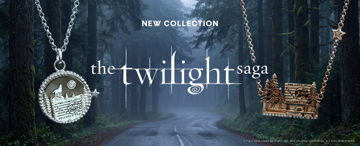 Promotional banner for The Twilight Saga New Collection featuring two pendants on chains: a silver pendant with a moonlit forest and cottage design, and a gold pendant with a cabin and trees. The background is a misty forest road with large trees, and the text 'NEW COLLECTION' and 'the twilight saga' appears across the center.