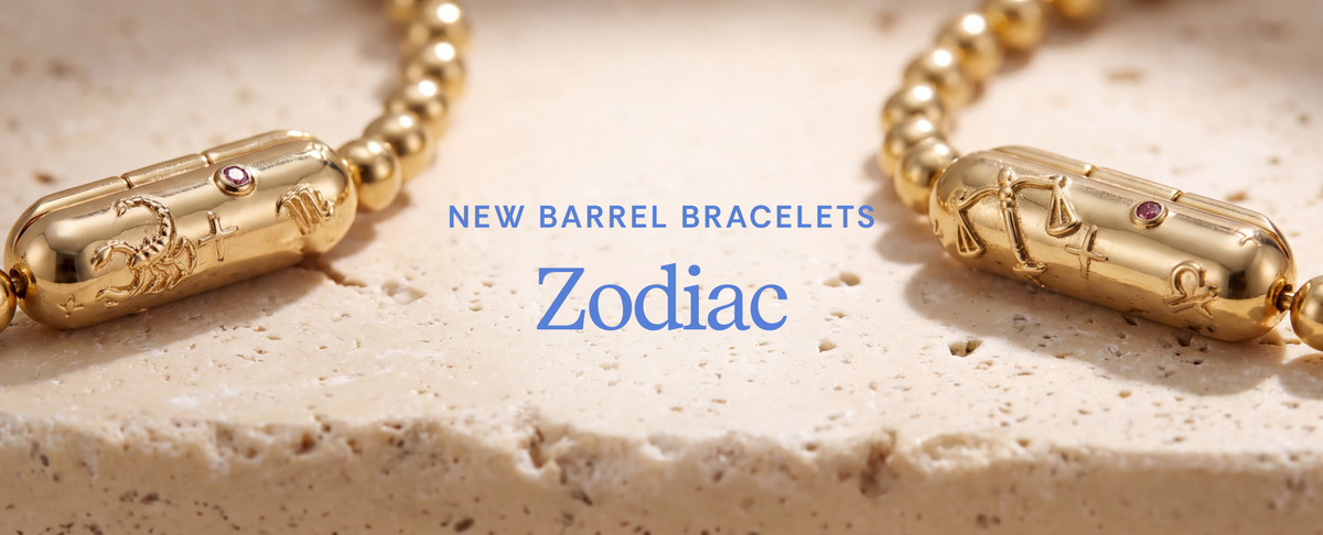 Gold-toned barrel bracelets with engraved zodiac symbols and small pink gemstone accents, displayed on a light stone surface with blue text reading 'NEW BARREL BRACELETS Zodiac'.