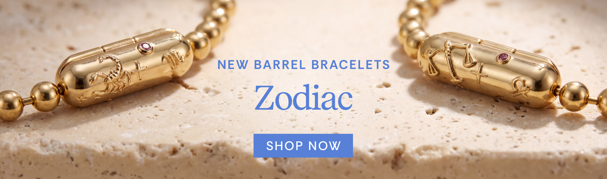 Gold-toned barrel-shaped zodiac bracelet charms displayed on a textured stone surface with promotional text reading 'New Barrel Bracelets Zodiac' and a blue 'Shop Now' button.