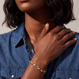 A close-up lifestyle shot of a model wearing a delicate gold chain bracelet with small gold charms and clear rhinestone accents, paired with a blue denim shirt.