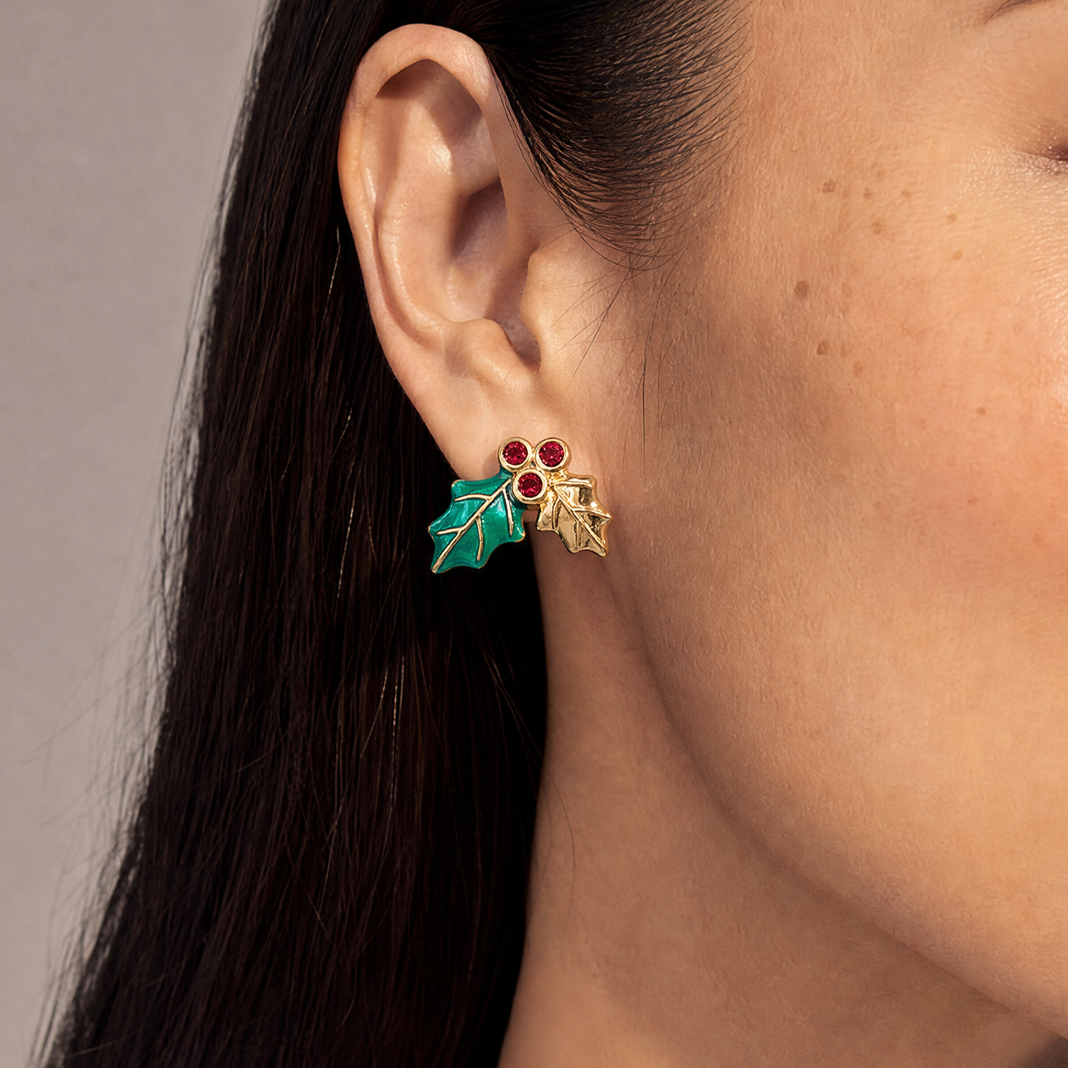 A close-up of a model's ear wearing a pair of gold holly leaf stud earrings with red berries and teal enamel details, set against a neutral gray background.
