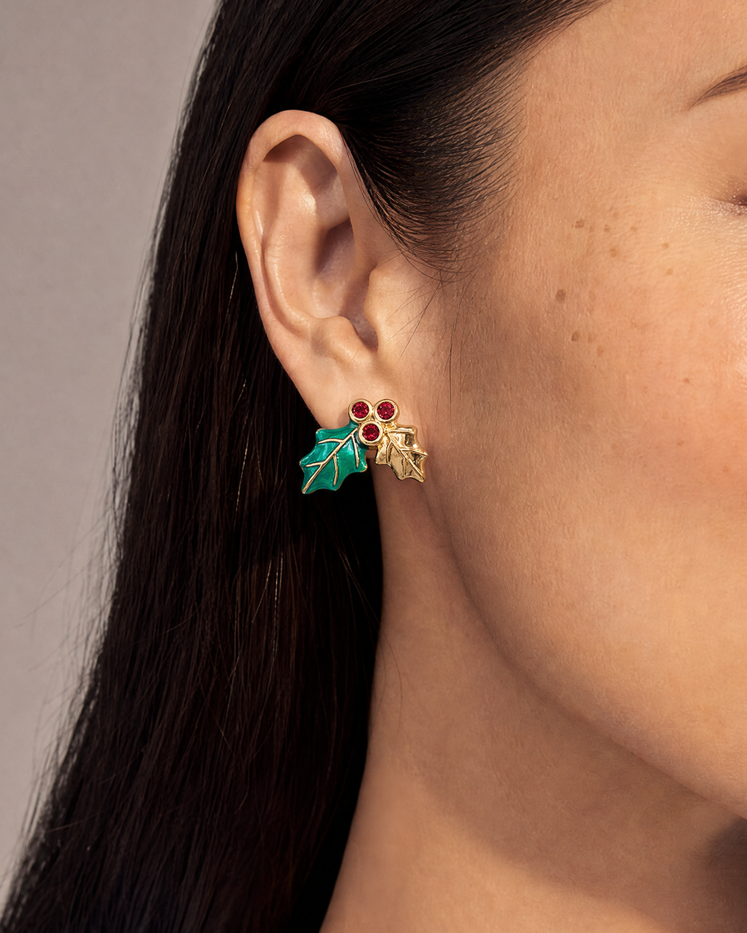 A close-up of a model's ear wearing a pair of gold holly leaf stud earrings with red berries and teal enamel details, set against a neutral gray background.
