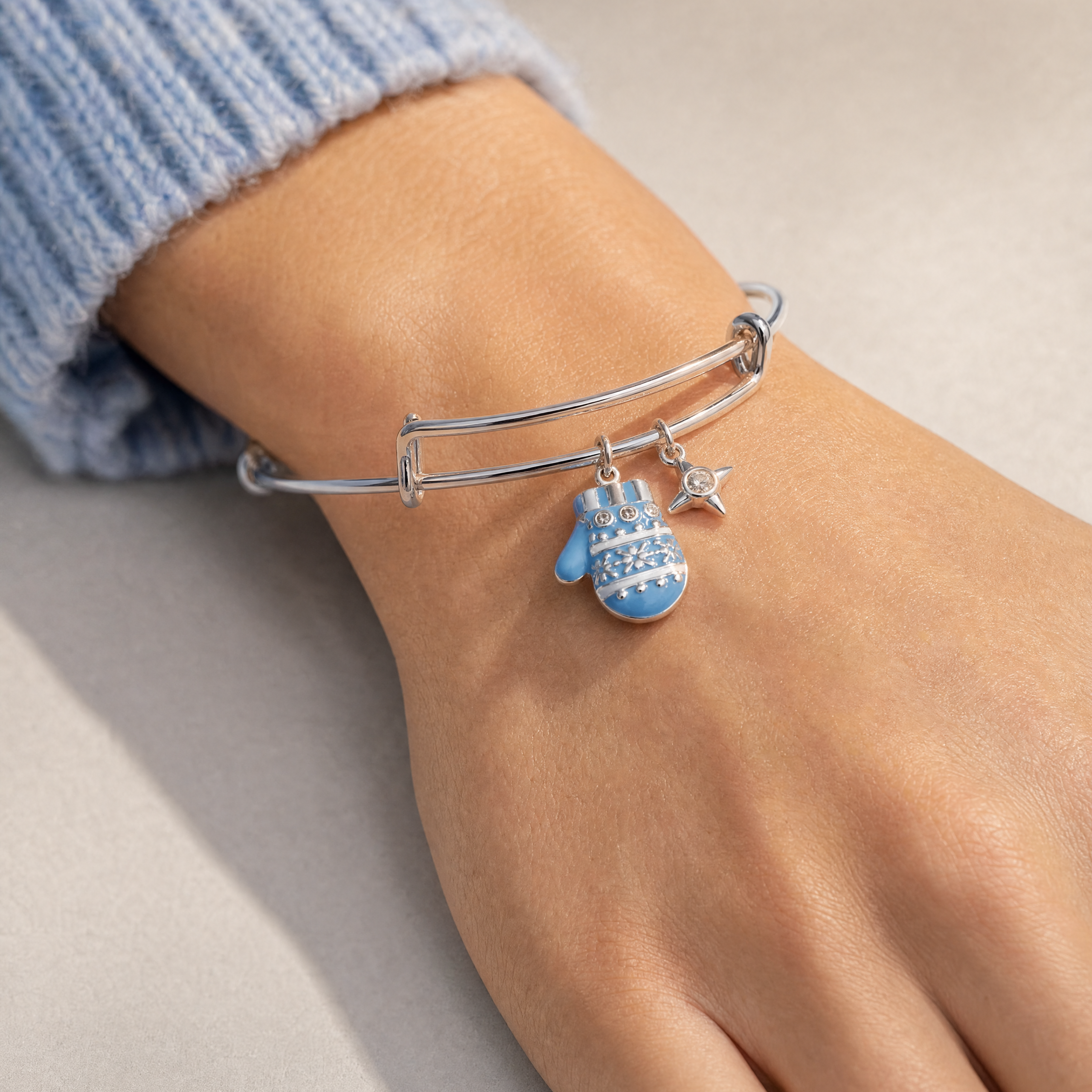 A close-up of a person's wrist wearing a silver bangle bracelet with a blue patterned mitten charm and a small silver star charm, paired with a light blue ribbed sweater cuff.