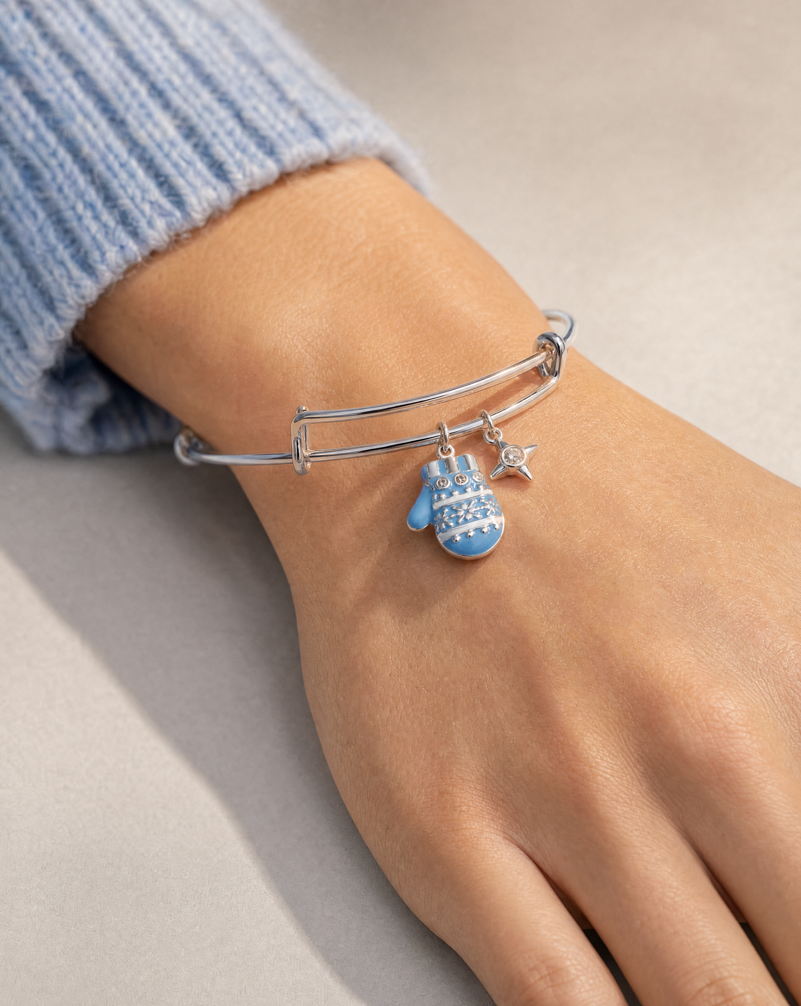 A close-up of a person's wrist wearing a silver bangle bracelet with a blue patterned mitten charm and a small silver star charm, paired with a light blue ribbed sweater cuff.