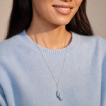 A close-up of a woman wearing a delicate silver necklace with a small blue and white striped shell pendant, paired with a light blue knit sweater.