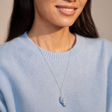 A close-up of a woman wearing a delicate silver necklace with a small blue and white striped shell pendant, paired with a light blue knit sweater.
