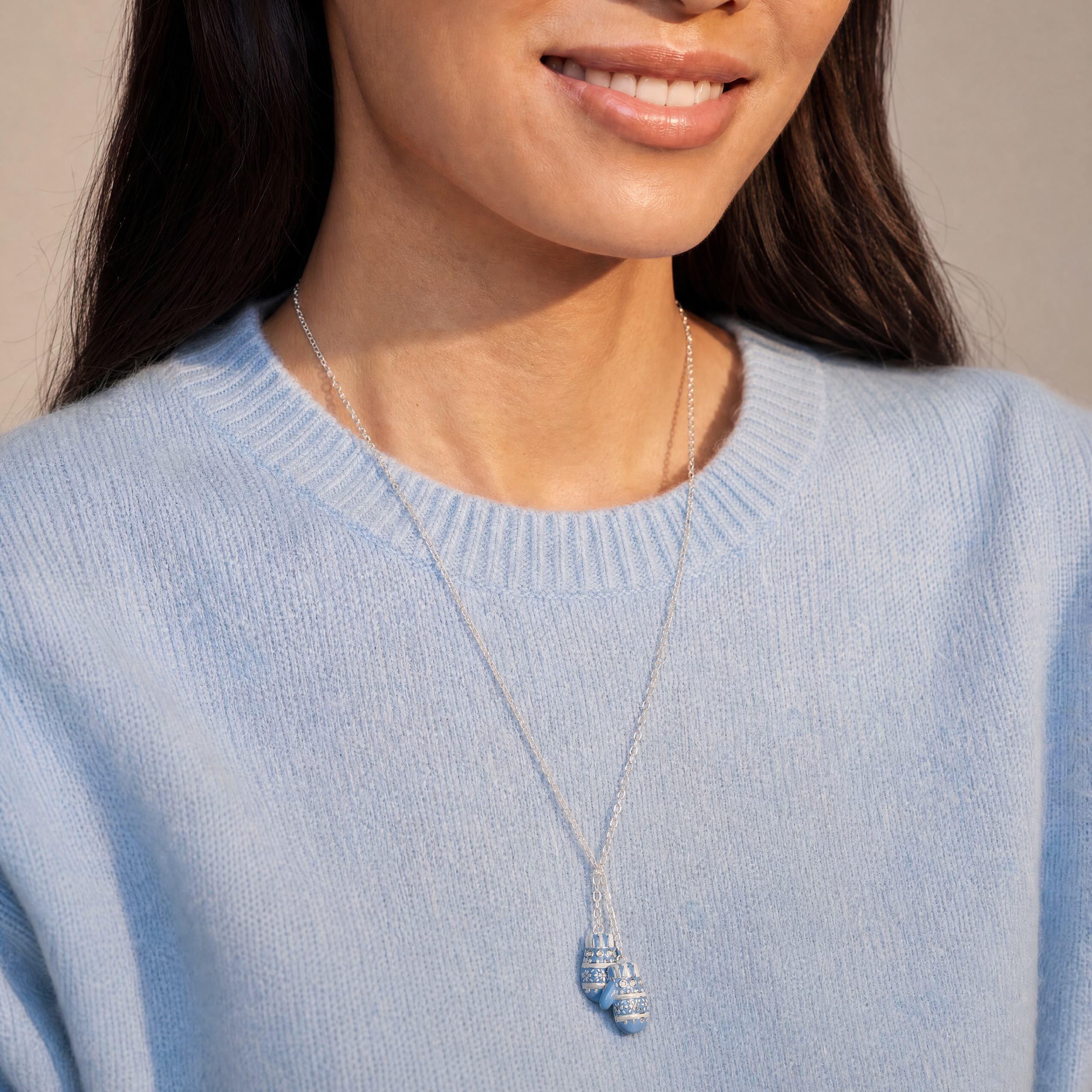 A close-up of a woman wearing a delicate silver necklace with a small blue and white striped shell pendant, paired with a light blue knit sweater.