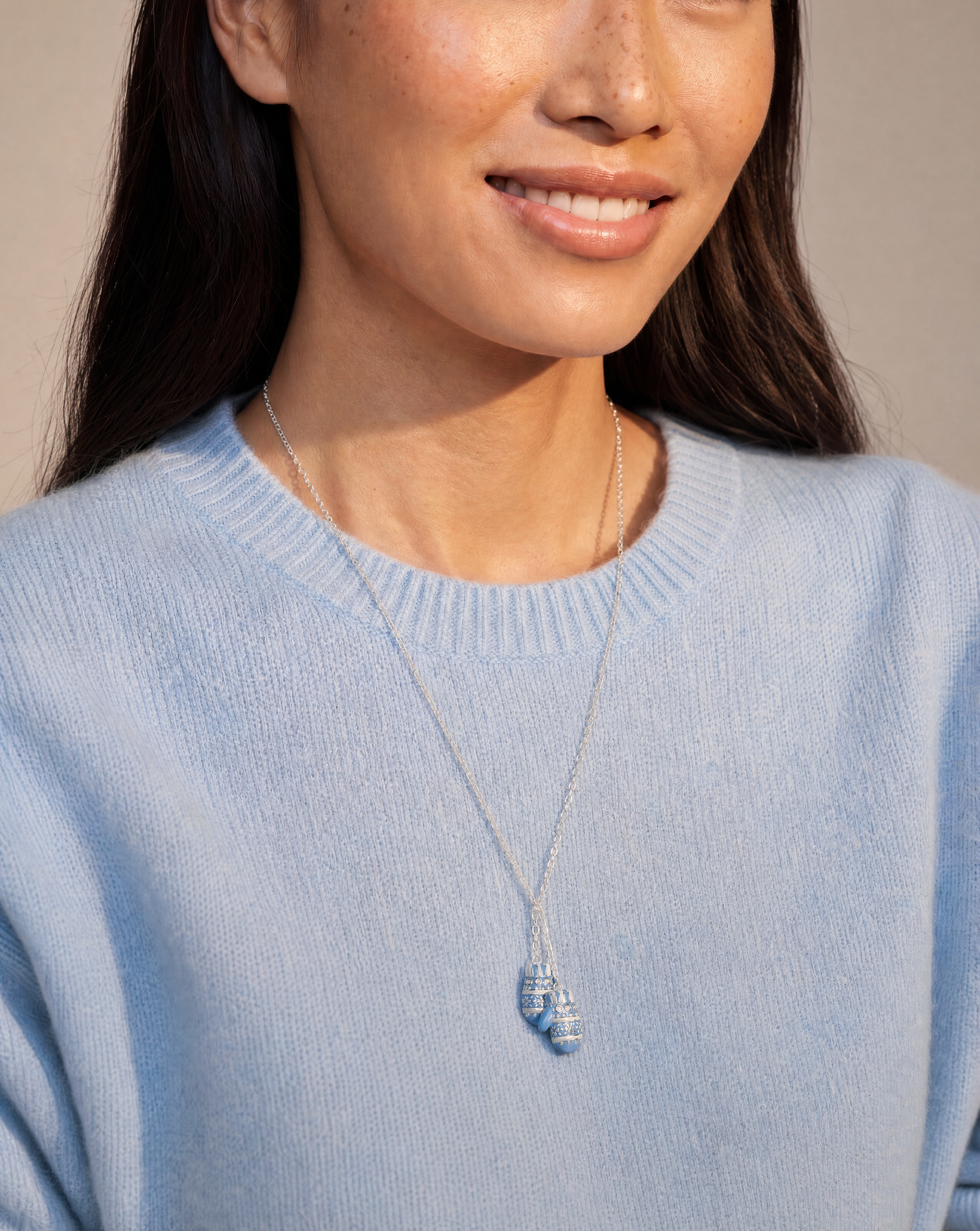 A close-up of a woman wearing a delicate silver necklace with a small blue and white striped shell pendant, paired with a light blue knit sweater.