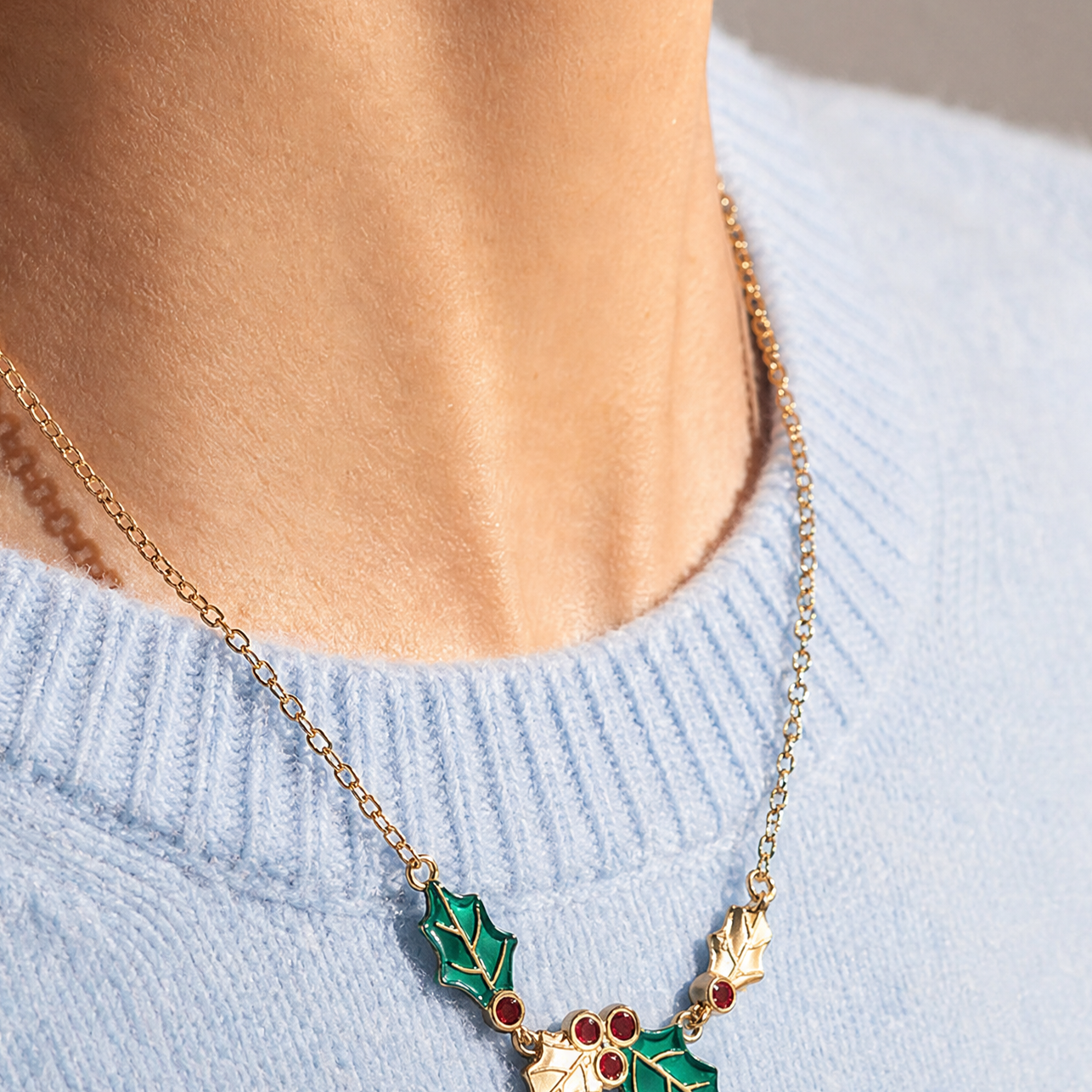 A gold chain necklace with a holly leaf pendant featuring green enamel and small red berry details, worn by a model in a light blue knit sweater.