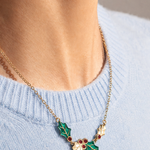 A gold chain necklace with a holly leaf pendant featuring green enamel and small red berry details, worn by a model in a light blue knit sweater.