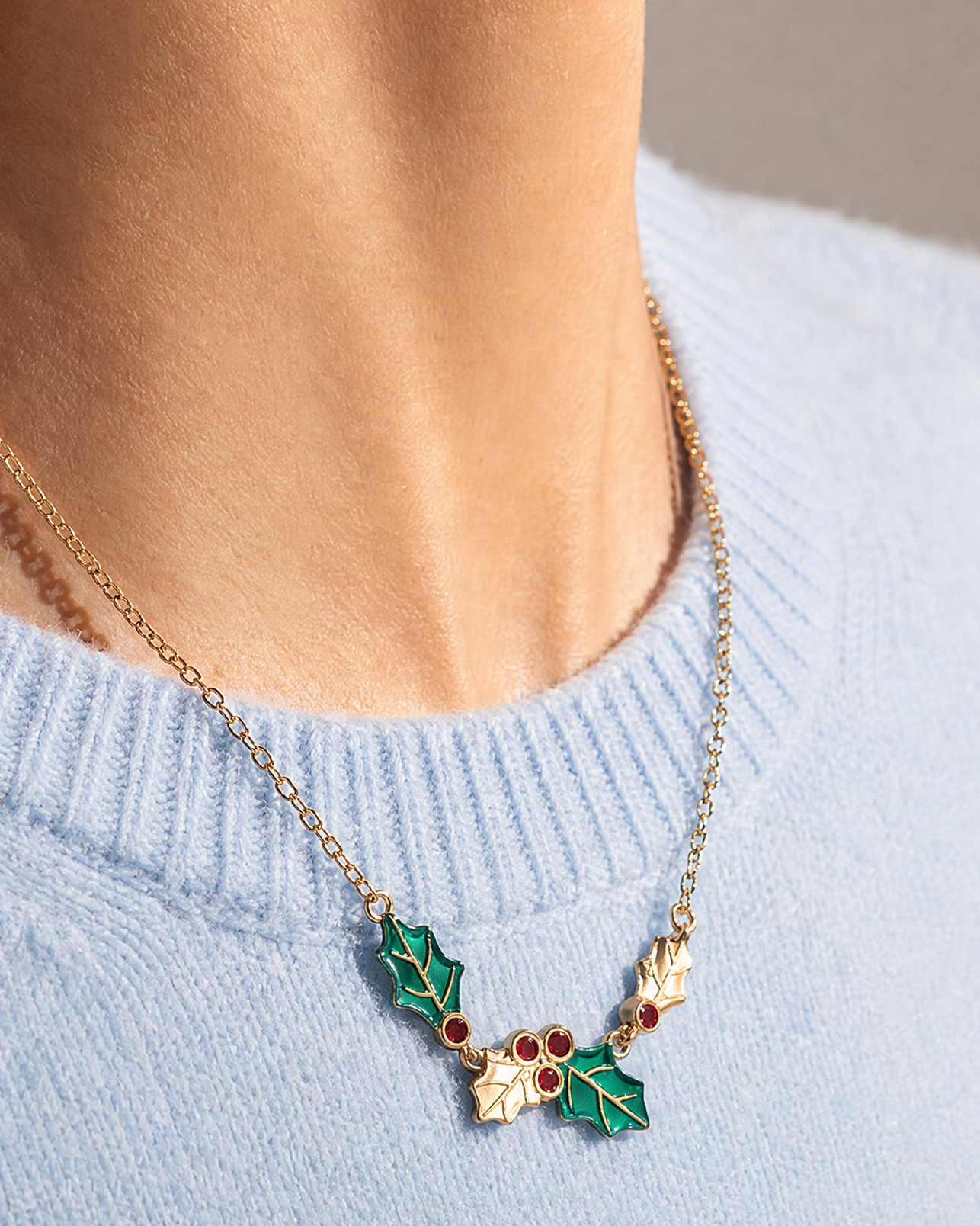 A gold chain necklace with a holly leaf pendant featuring green enamel and small red berry details, worn by a model in a light blue knit sweater.