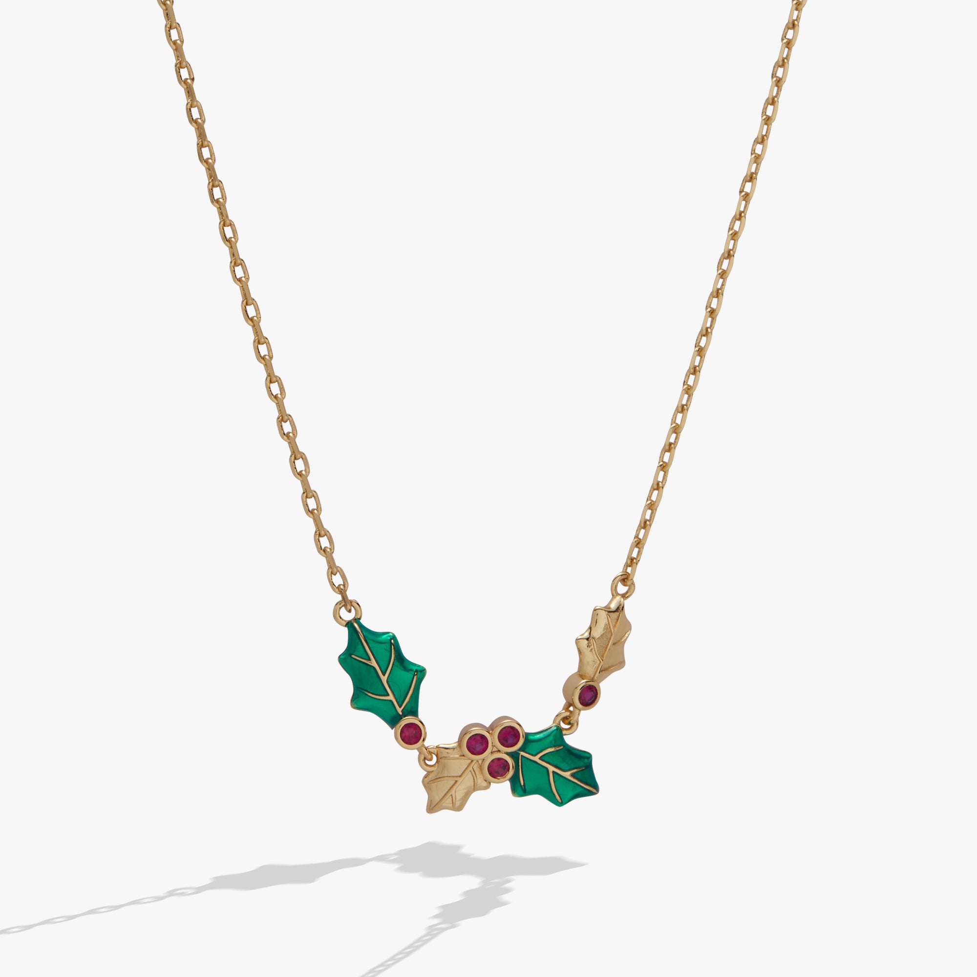 A gold-tone chain necklace featuring a holly pendant with green enamel leaves and small red gemstone accents, shown against a light gray background.